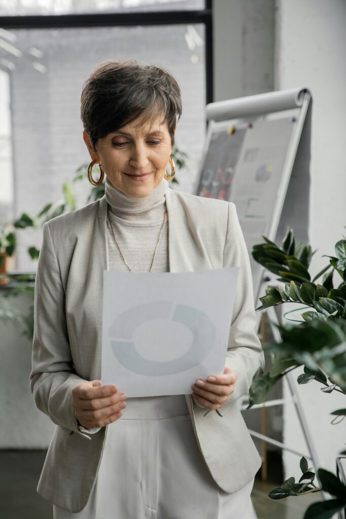 smiling and stylish mature woman, corporate manager, looking at document with analytics in office